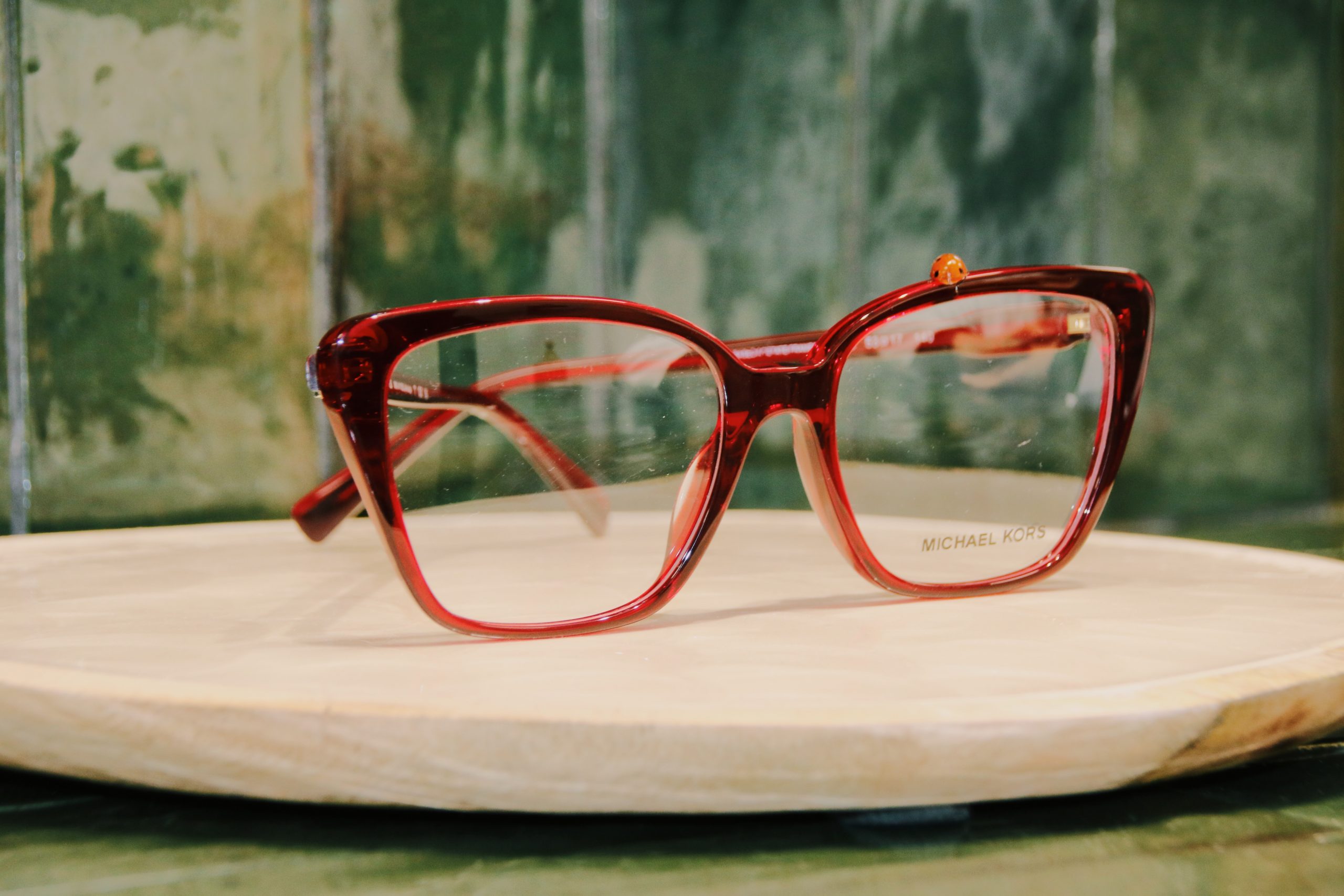 Foto de gafas rojas Michael Kors con una mariquita encima y de fondo columna de azulejos verde oliva en el local de Cámara Centro Óptico y Auditivo Montecarmelo.